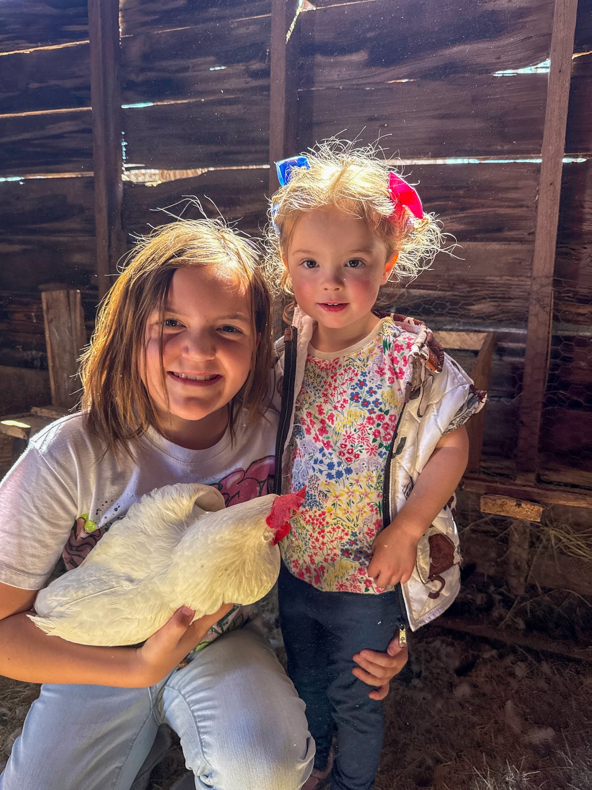 Two children smiling with a white chicken inside a rustic coop